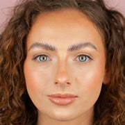 Close-up of a person with curly brown hair, light blue eyes, and a septum piercing looking directly at the camera. With distinct brows and Boldface Lashes - Short and Sweet, they're wearing cruelty-free natural makeup and a soft pink lip color.